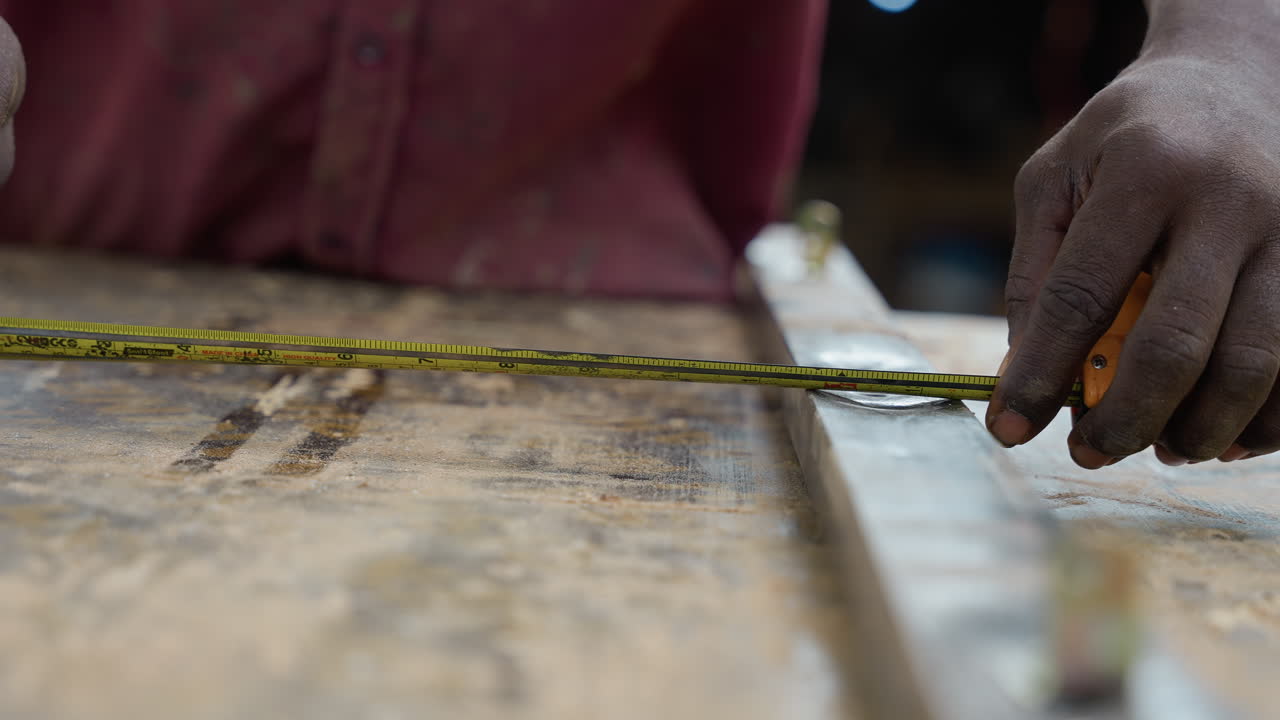 Primo piano di mani che misurano il legno su un banco da lavoro. Un falegname esperto usa un metro a nastro e una riga per segnare una linea di taglio precisa su una tavola usurata, granelli di segatura su una superficie ruvida, una guida metallica arrugginita, atmosfera concentrata.