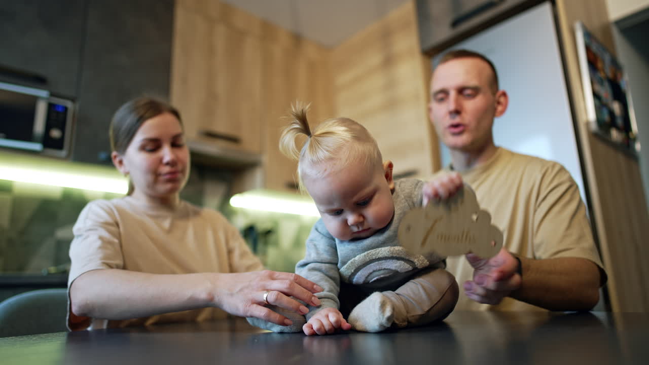 Cute baby boy with a tail on his blond hair sits on the table. Loving parents are near their adorable child. Kitchen backdrop.