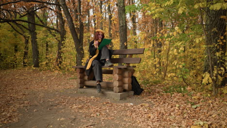 female artist sketching seated on wooden park bench with bag on floor in solace surrounded by golden autumn trees in peaceful outdoor setting, immersed in creativity, wearing yellow beret and scarf