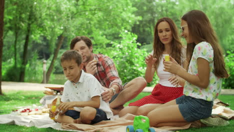 Siblings-playing-with-soap-bubbles-in-park.-Family-sitting-on-blanket-at-picnic