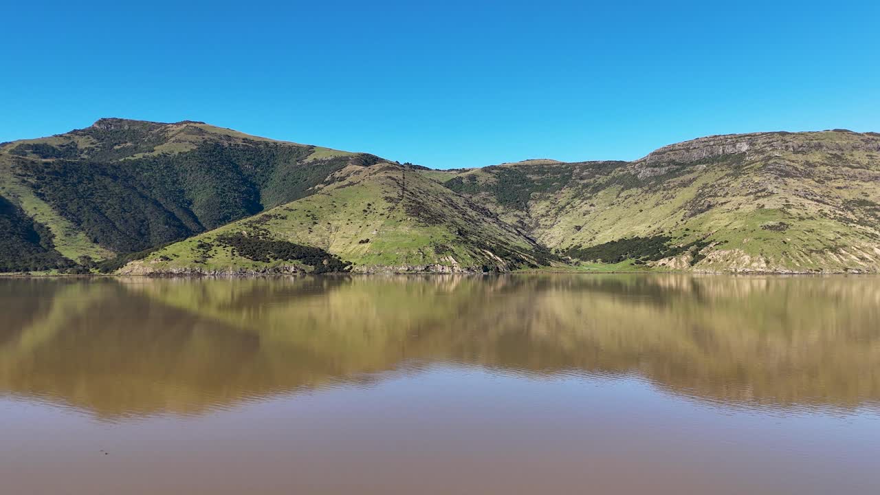Aerial footage captures Akaroa's serene landscape with lush hills and a reflective lake under clear blue skies