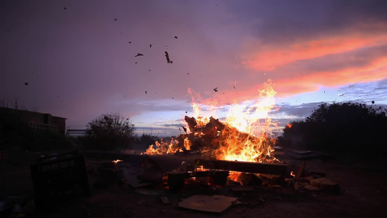 Old wooden items are burning in large open fire in a backyard. Sun is setting and pink and blue sky looks beautiful. Ash is flying everywhere; large piece of ash separates and flyes up. Footage in HD