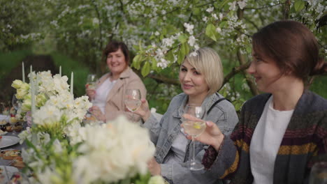 Fröhliche-Menschen-Speisen-Im-Garten,-Trinken-Wein-Und-Cocktails-Und-Sitzen-An-Einem-Mit-Blumen-Geschmückten-Tisch