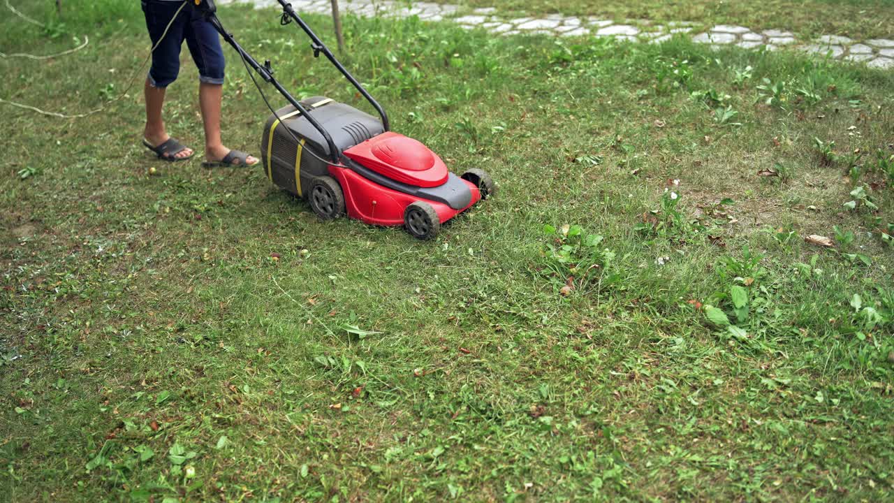 Electric lawn mower in the garden. Boy cutting grass by lawn mowing machine in summer day. Gardening activity.