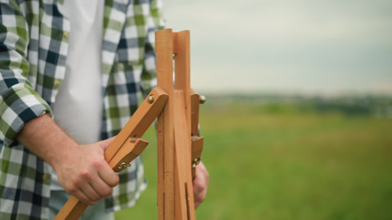 A person in a plaid shirt is holding and setting up a wooden tripod outdoors in a grassy field. The close-up shot focuses on the hands and tripod, process of preparation in a natural, serene setting