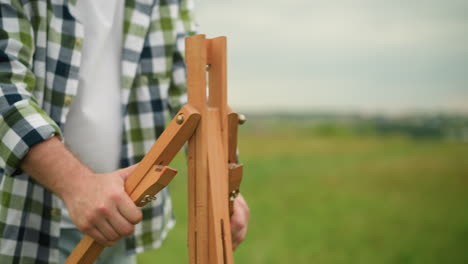 a person in a plaid shirt is holding and setting up a wooden tripod outdoors in a grassy field. the close-up shot focuses on the hands and tripod, process of preparation in a natural, serene setting