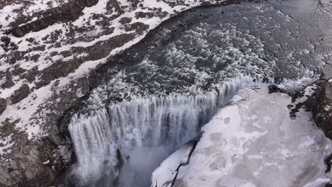 aerial of dettifoss waterfall in iceland crashing into ice with powerful mist and snowy cliffs