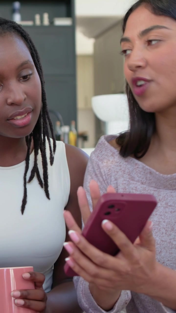 Vertical video: Leaning mauve-white pair as right woman tapping maroon phone on home couch sharing
