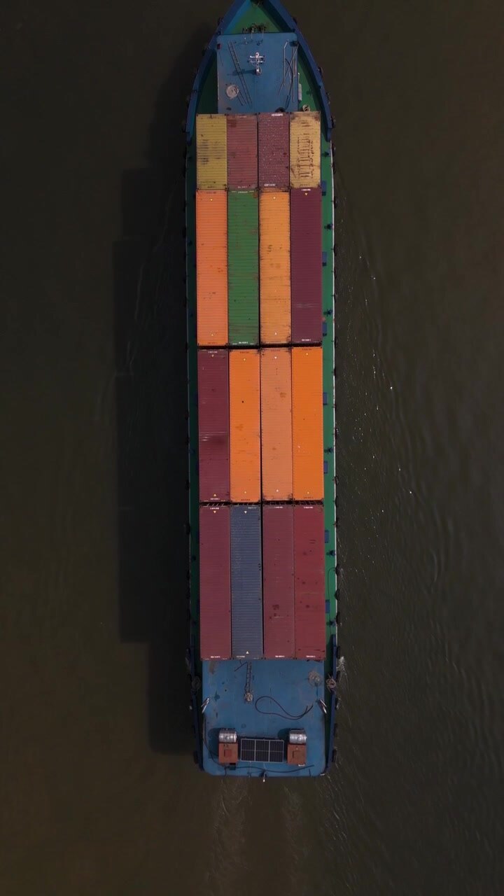 Colorful shipping containers on a river freighter from top down aerial view in dramatic light. Colors are orange, , red and blue. Water is a dark shade. Vertical format.