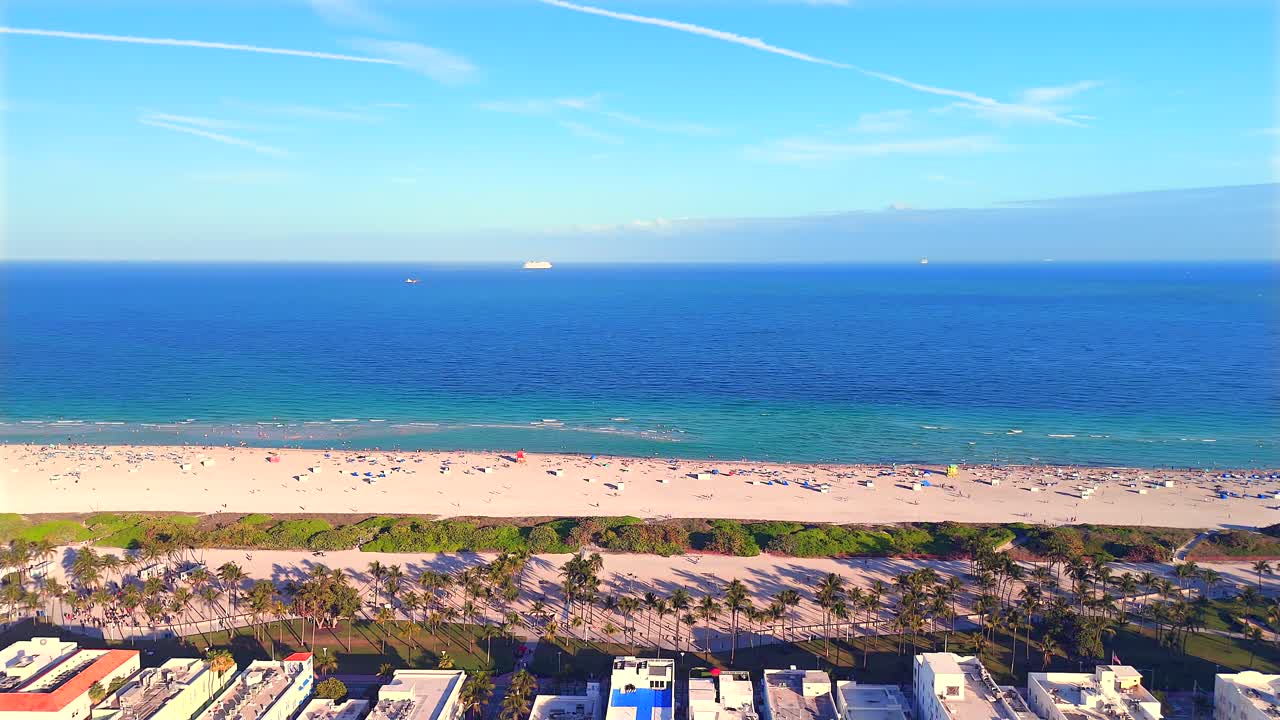 Drone captures a stunning 360° view over South Beach, Miami, revealing ocean waves, golden sand, South Pointe, the downtown skyline at sunset, and the coastline stretching north.