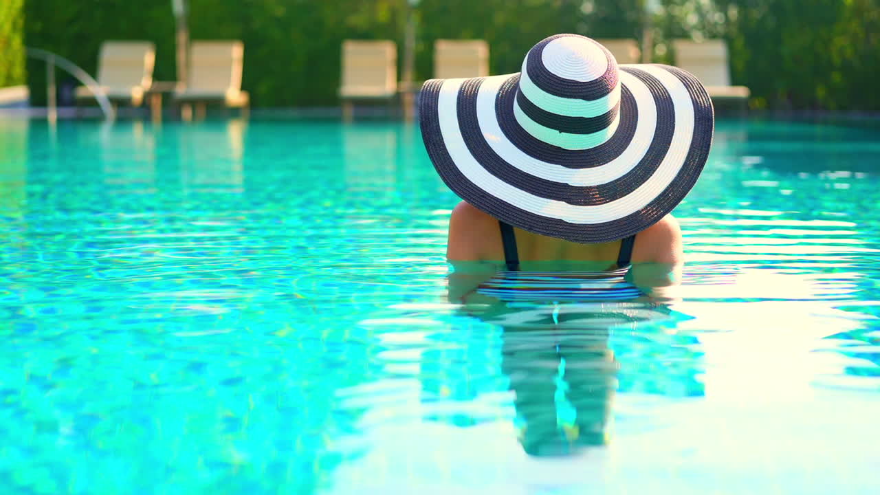Rückenansicht einer Frau mit Sommerhut und Badeanzug, die an einem sonnigen Tag in einem Schwimmbecken steht, Ganzaufnahme.