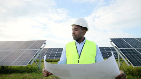 vista frontal de un joven ingeniero afroamericano sosteniendo un plan solar y mirando alrededor en una plantación solar