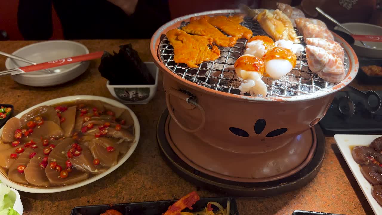 A person prepares and grills assorted seafood and squid on a tabletop charcoal barbecue in a warmly lit restaurant, with close-up camera angles and minimal movement