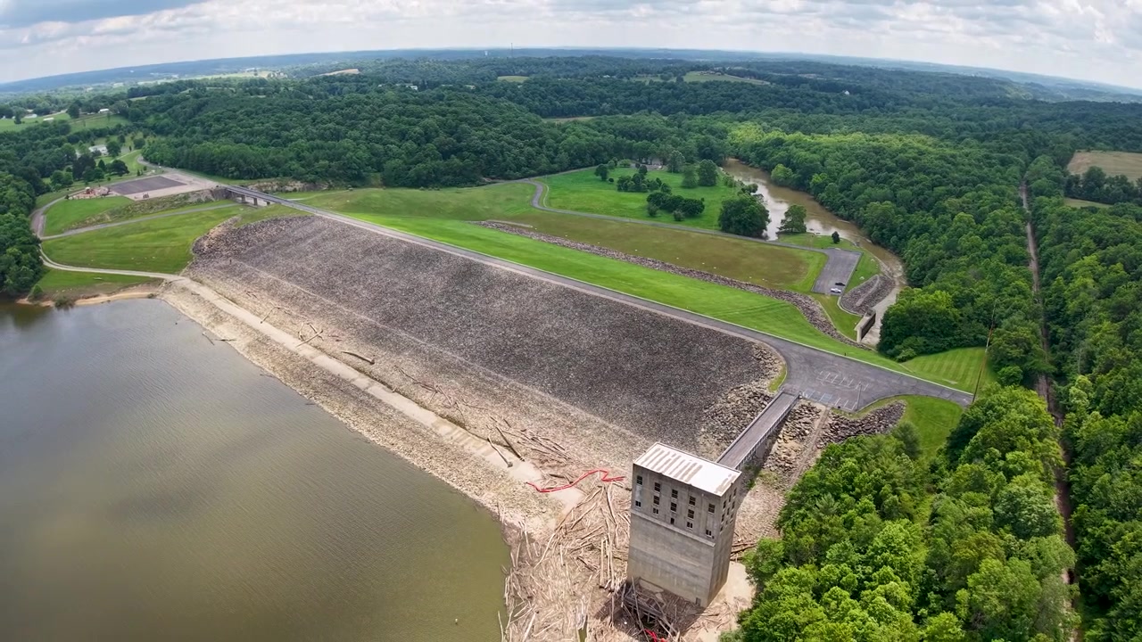 Aerial view of a concrete dam and spillway beside calm reservoir water and dense green woodland