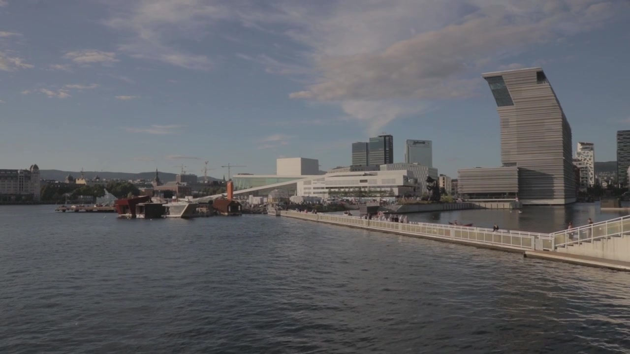 Weitwinkelaufnahme der Bjørvika-Uferpromenade in Oslo, Norwegen, mit dem modernen Munch-Museum, dem Opernhaus Oslo und schwimmenden Saunen entlang des Oslofjords unter blauem Himmel an einem ruhigen Sommertag
