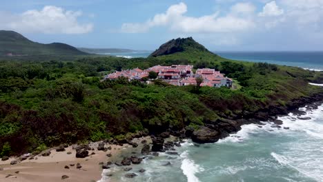 Coastal-resort-town-with-red-roofed-buildings-nestled-by-the-sea,-lush-greenery,-sunny-day,-aerial-view