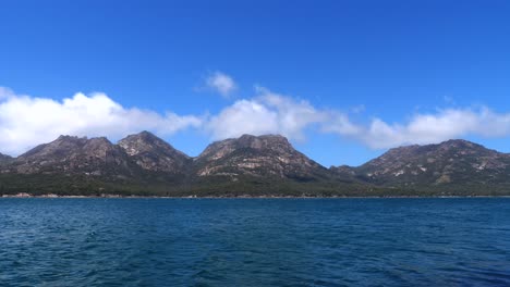 在晴朗的晴朗的夏天,白云在岩石山脉上滚动,前景是蓝色的海洋水,弗雷西内特,塔斯马尼亚