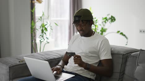 Young-black-man-holds-plastic-bank-card,-sitting-on-a-comfortable-sofa,-shopping-using-a-laptop