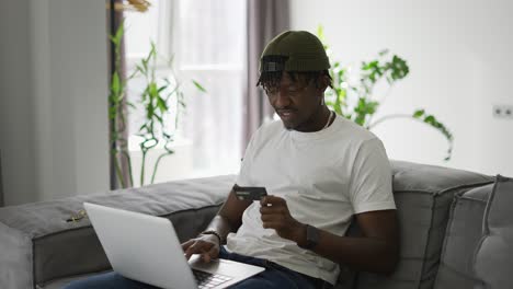 young black man holds plastic bank card, sitting on a comfortable sofa, shopping using a laptop