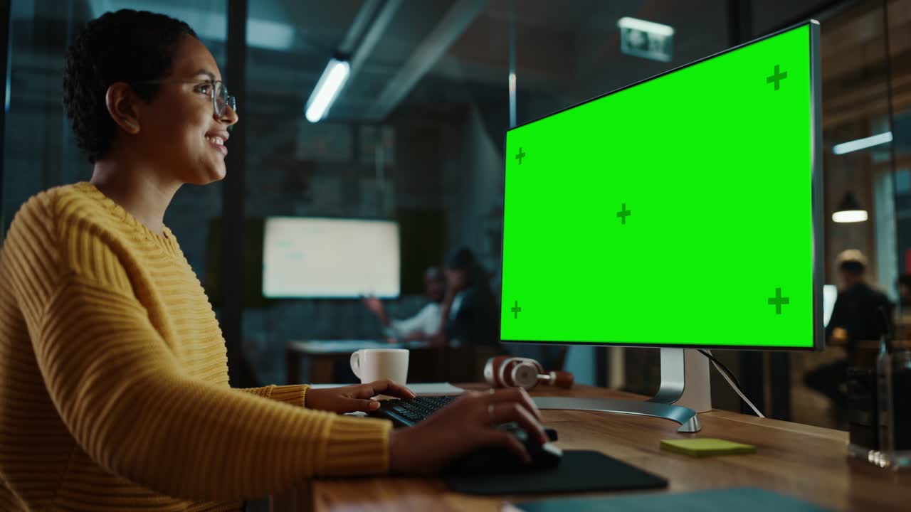 joven especialista multiétnico trabajando en una computadora de escritorio con pantalla verde simulacro de visualización en la oficina creativa. hermosa gerente femenina diversa con cabello corto y gafas está usando un jumper amarillo.