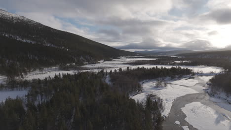 backwards flying droneshot over a snowy valley in norway with clouds in the sky and a low hanging sun near sunrise with trees and rivers underneath log