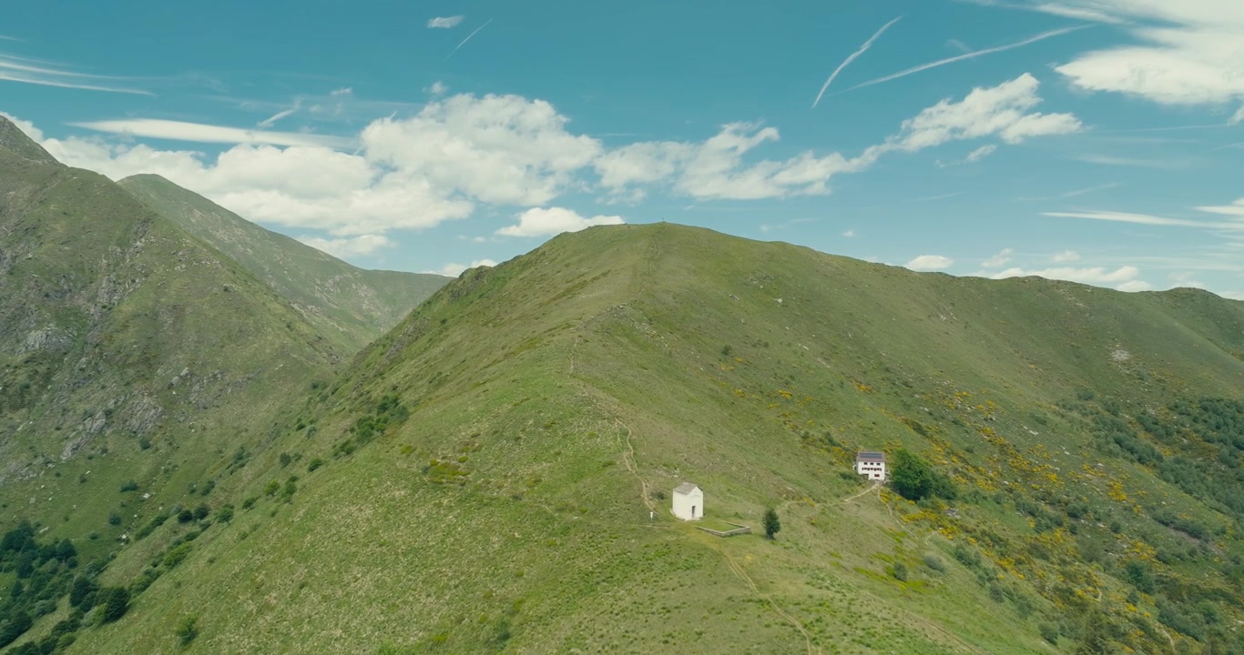 Drone flyver over en frodig bjergryg mod et topkors, der afslører lag af alpetoppe under en klar blå sommerhimmel. Betagende luftfoto af vandring og bjerglandskab.