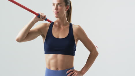 Una-Joven-Atleta-Caucásica-Sostiene-Una-Jabalina-En-El-Gimnasio-Con-Un-Fondo-Blanco.