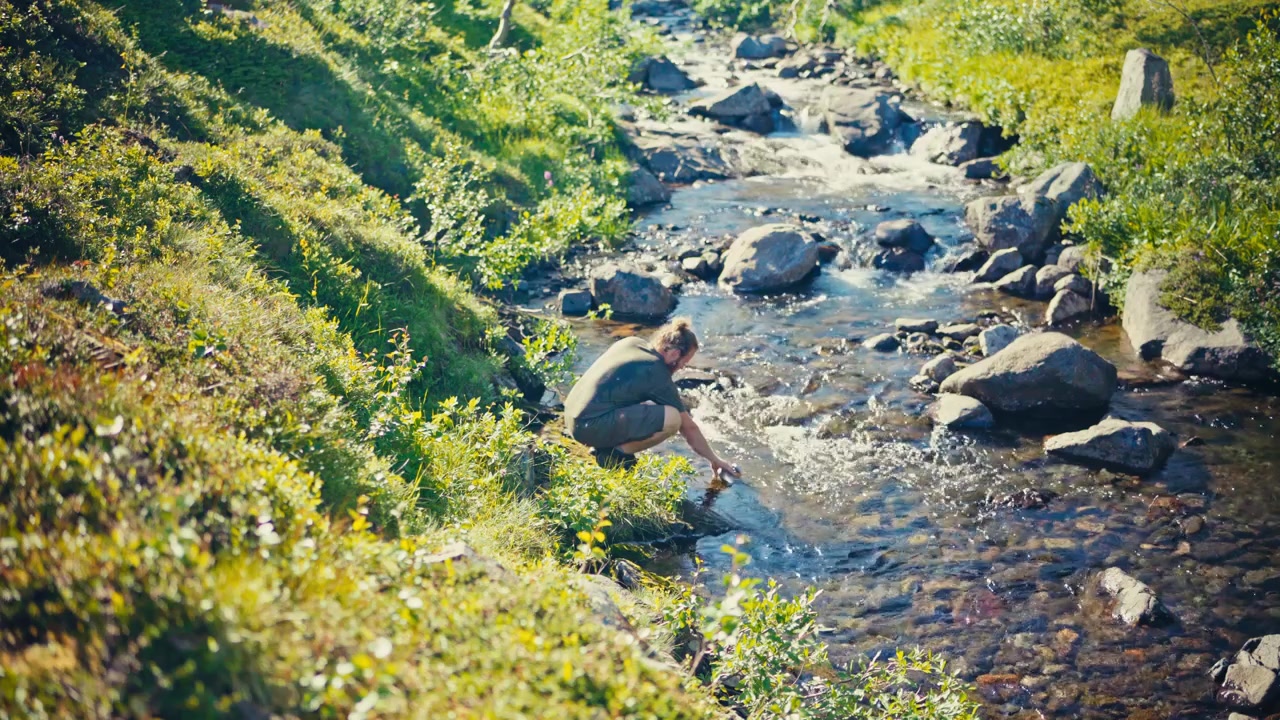 Pria Norwegia dengan Tumbler Mengambil Air dari Sungai Berbatu yang Mengalir. Rekaman Sudut Tinggi.