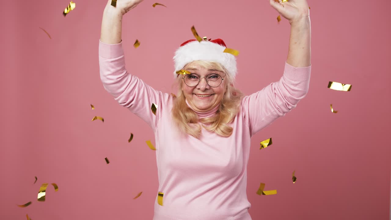 Atmosfera natalizia. Anziana signora felice con cappello da Babbo Natale che lancia glitter dorati e balla, sfondo rosa, slow motion.