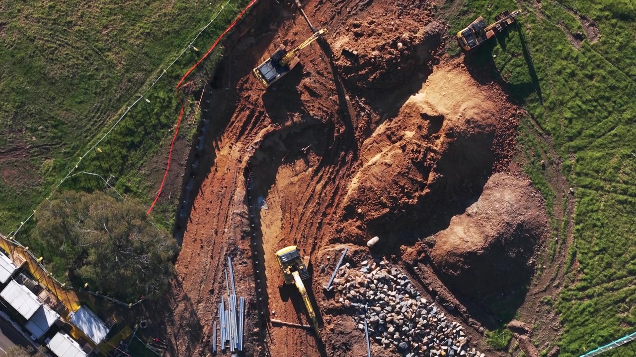 Drone video captures a South East Melbourne construction site with excavators, trucks, and steel beams. Sunlit earthworks, green trees, and suburban rooftops create a dynamic, industrial scene.