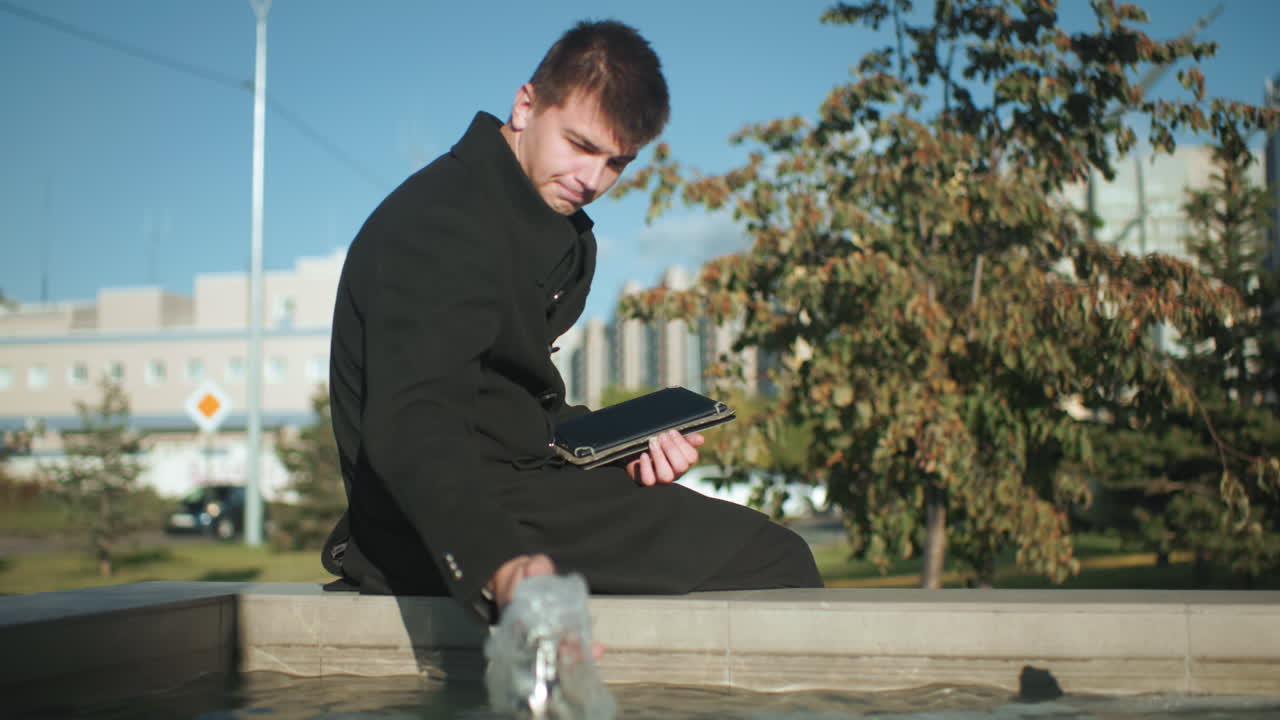 물 에 앉아있는 남자가 타블을 사용하면서 물 표면을 만지고 다리에 앉아 있으며, 도시 초록색, 현대적인 건물, 그리고 밝은 빛 하늘 아래 움직이는 자동차로 둘러싸여 있습니다.