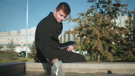 Man seated beside spring water touching water surface while using tablet resting on his legs, surrounded by urban greenery, modern buildings, and moving cars under bright sunny skies