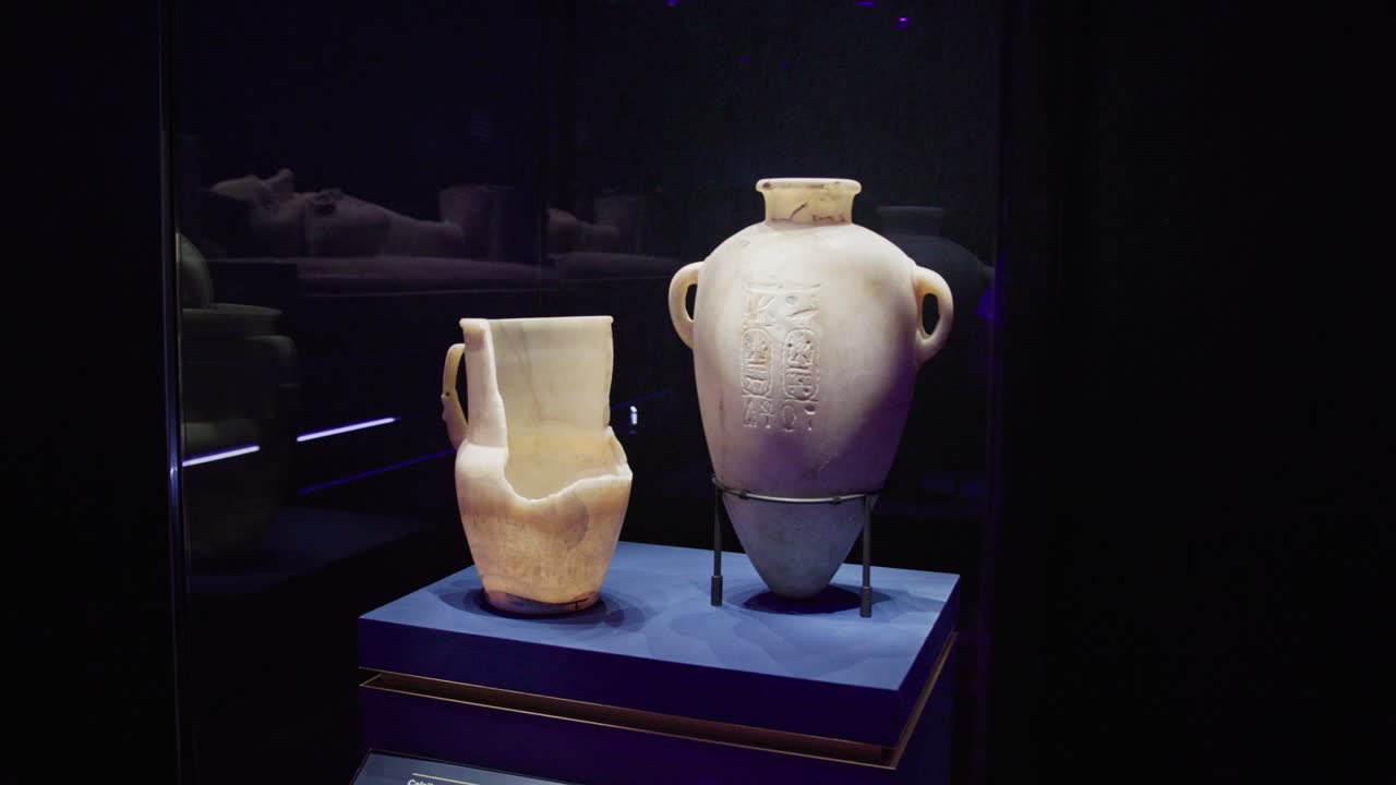 Vasijas de alabastro del Antiguo Egipto, incluyendo un vaso canopo, expuestas en una vitrina de museo tenuemente iluminada con inscripciones y soportes de conservación.