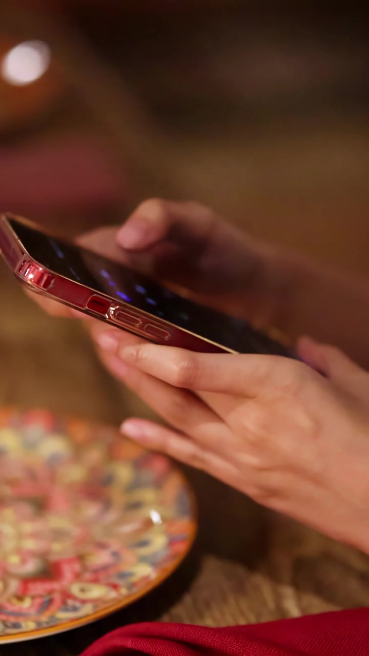 Mani femminili interagiscono con uno smartphone rosso sopra un piatto decorativo in un ambiente di ristorante illuminato caldamente, ripreso in un primo piano stabile.