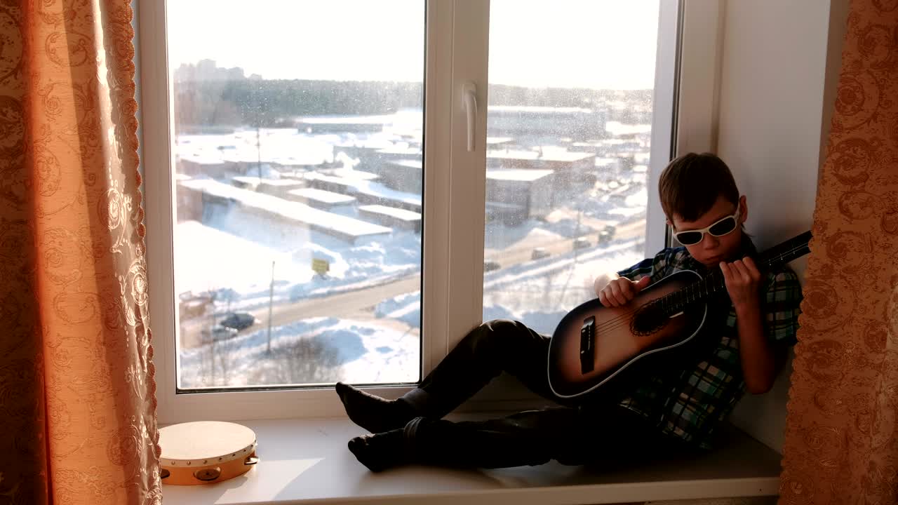 un niño con gafas de sol toca la guitarra sentado en el alféizar de la ventana.