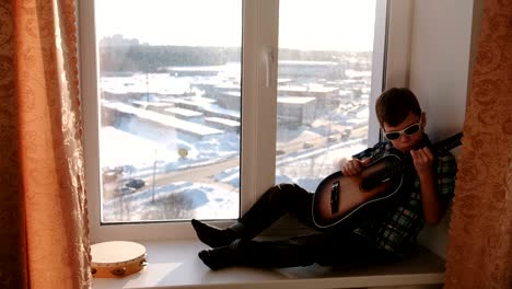 playing a musical instrument. boy in sunglasses plays the guitar sitting on the windowsill.