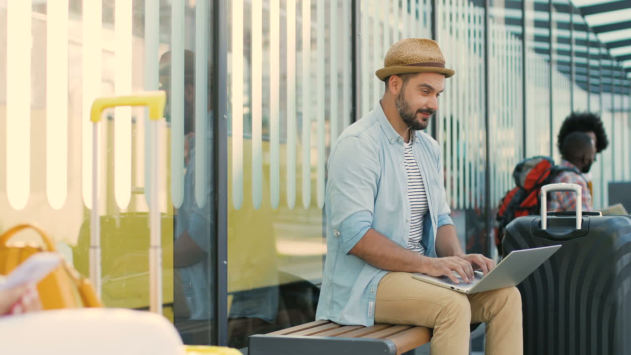 joven viajero caucásico guapo con sombrero sentado en un banco en la parada de autobús con maleta y esperando el transporte mientras usa una computadora portátil