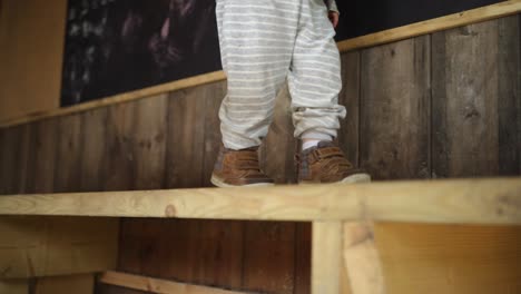 toddler boy slowly walks right along wooden bench in hut, mid shot feet