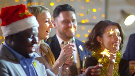 happy women and men holding sparklers and glasses of champagne and dancing at christmas party as golden confetti falling on them from above