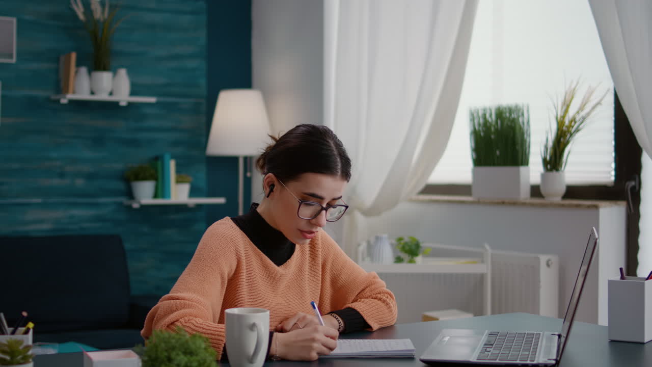 Young adult attending video call meeting on laptop and taking notes