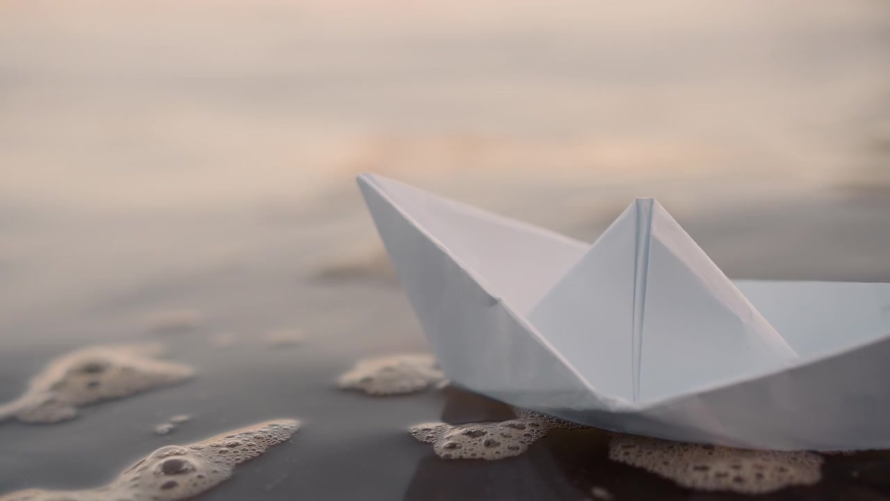 Una barchetta di carta bianca galleggia dolcemente su acqua calma con leggere increspature e bolle, muovendosi gradualmente in avanti in un sereno ambiente acquatico. La scena presenta luce naturale che si riflette sulla superficie bagnata