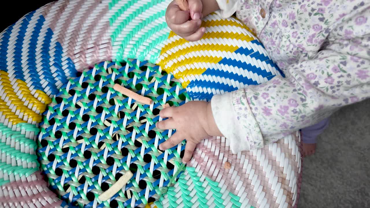 Baby hands reaching for healthy vegetable sticks on a vibrant woven stool from an overhead view
