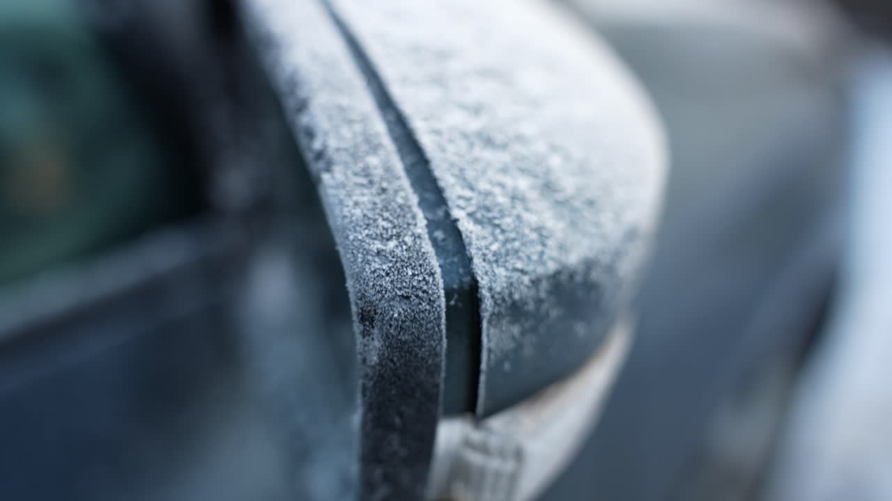 Makrokuva auton sivupeilistä, joka on kuuran ja jääkiteiden peitossa, ja jossa näkyy jäinen tekstuuri kylmänä talviaamuna pienellä syväterävyydellä.
