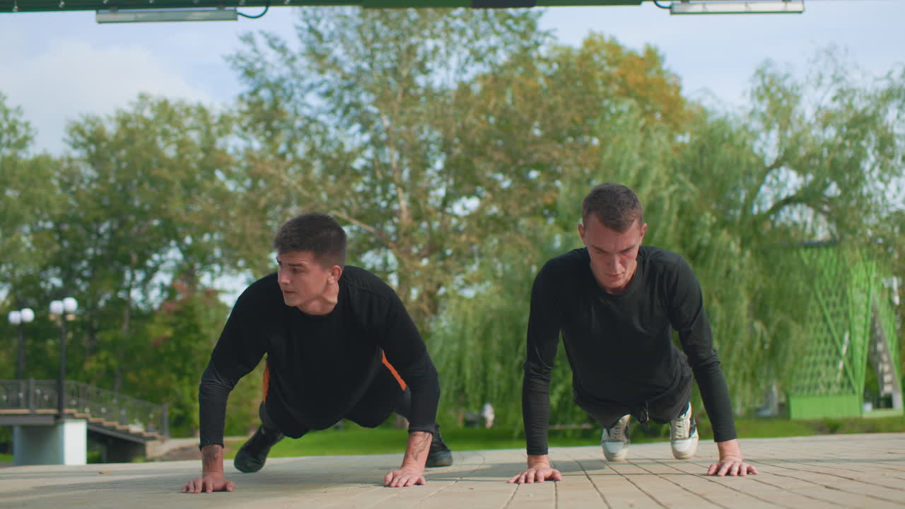 Due uomini fanno flessioni sotto un padiglione, sessione di fitness sincronizzata tra due uomini sotto un rifugio verde, due uomini impegnati in esercizi a corpo libero all'aperto con spirito competitivo e scenario autunnale