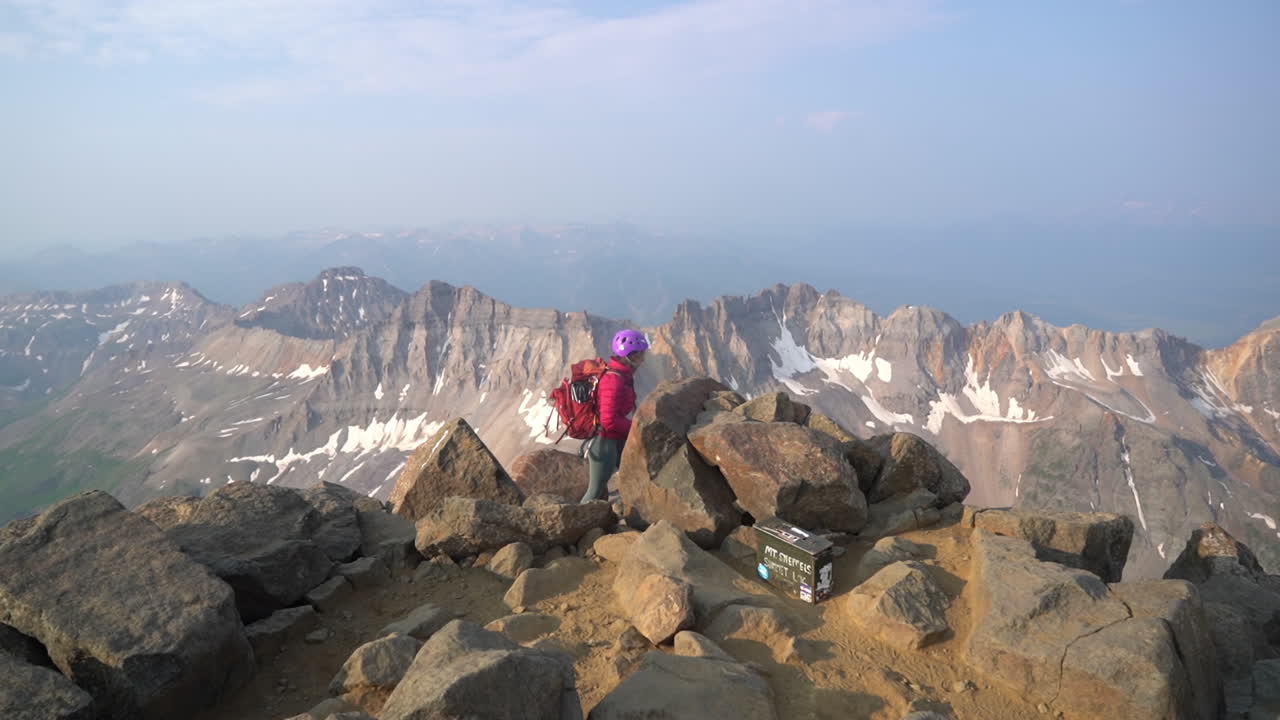 Donna solitaria sulla vetta del Monte Sneffels, escursionista con zaino e casco in cima con vista incredibile sulla catena montuosa, Colorado, USA.