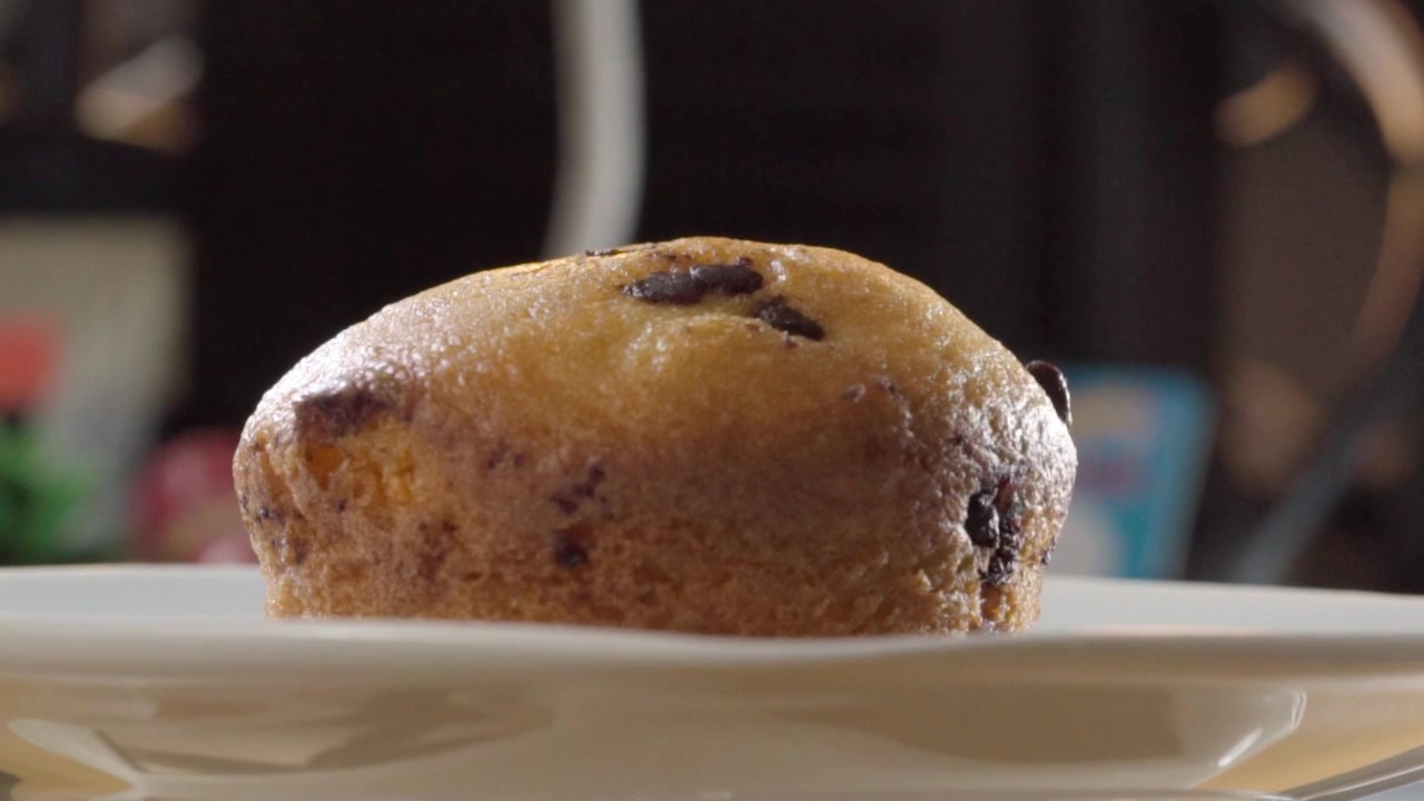 Muffin fresco con gocce di cioccolato appoggiato su un piatto di ceramica bianca, che mostra una superficie dorata e cotta, gocce di cioccolato fondente e una consistenza morbida in primo piano