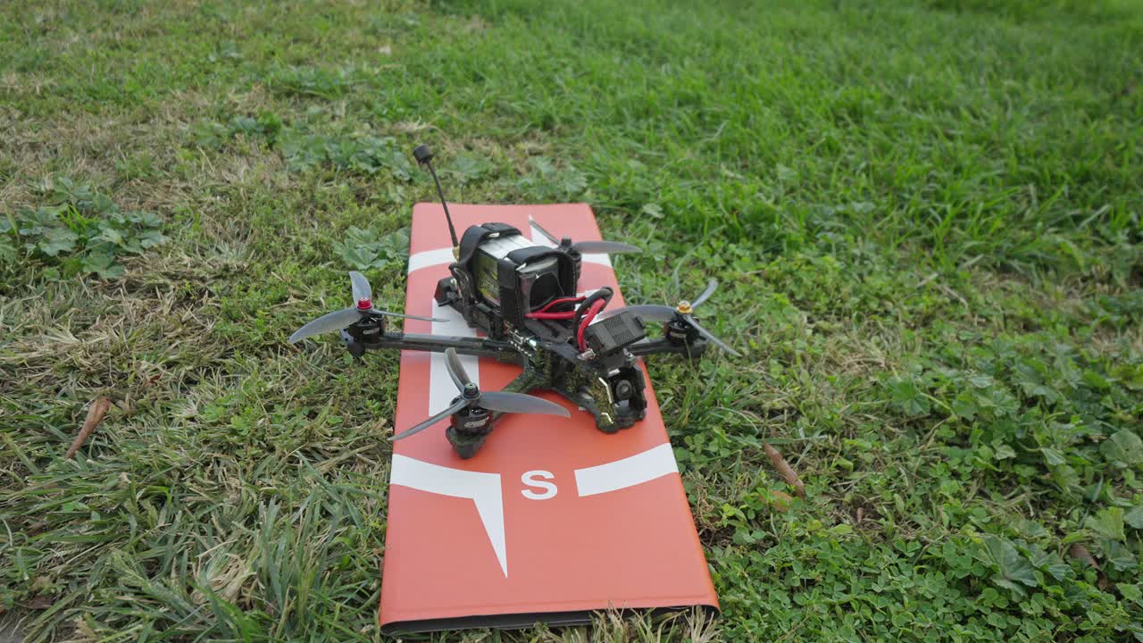 An FPV drone rests on a grass launch pad as the pilot switches it into arm mode, the motors giving a subtle twitch before liftoff