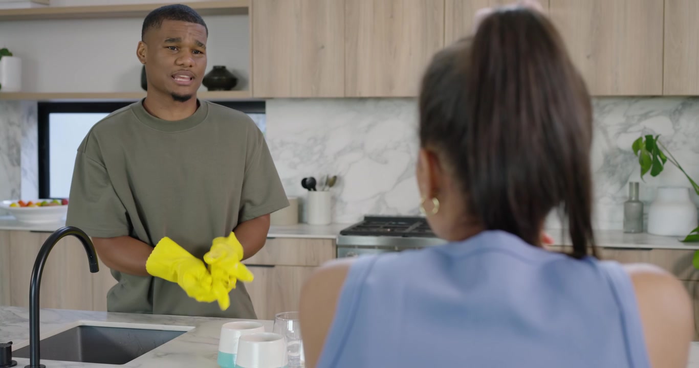 African American couple listening as man in yellow gloves washing dishes and explaining at island