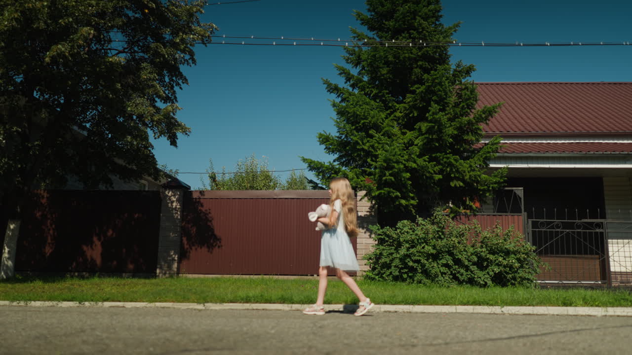 Side view of young girl walking beside pavement holding stuffed toy on calm residential street with background of green tree, fence, and house under clear blue sky