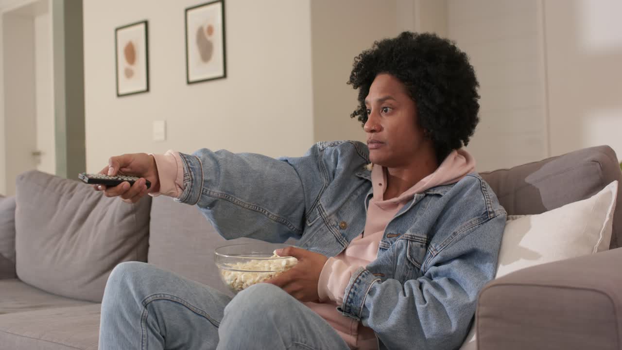 Afroamerikansk mand går ind i stuen, trykker på fjernbetjeningen og spiser popcorn, mens han ser tv på sofaen